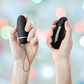 Two hands each hold a black electronic device against a blurred, colorful background. The left hand grips the small, oval B Swish Bnaughty Deluxe Unleashed with a loop, while the right hand holds a larger, oblong wireless remote with buttons—both featuring a waterproof design for added convenience.
